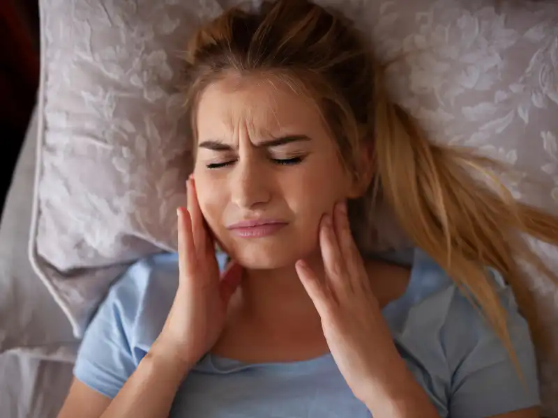 A woman lying in bed with a painful expression, clutching her jaw.